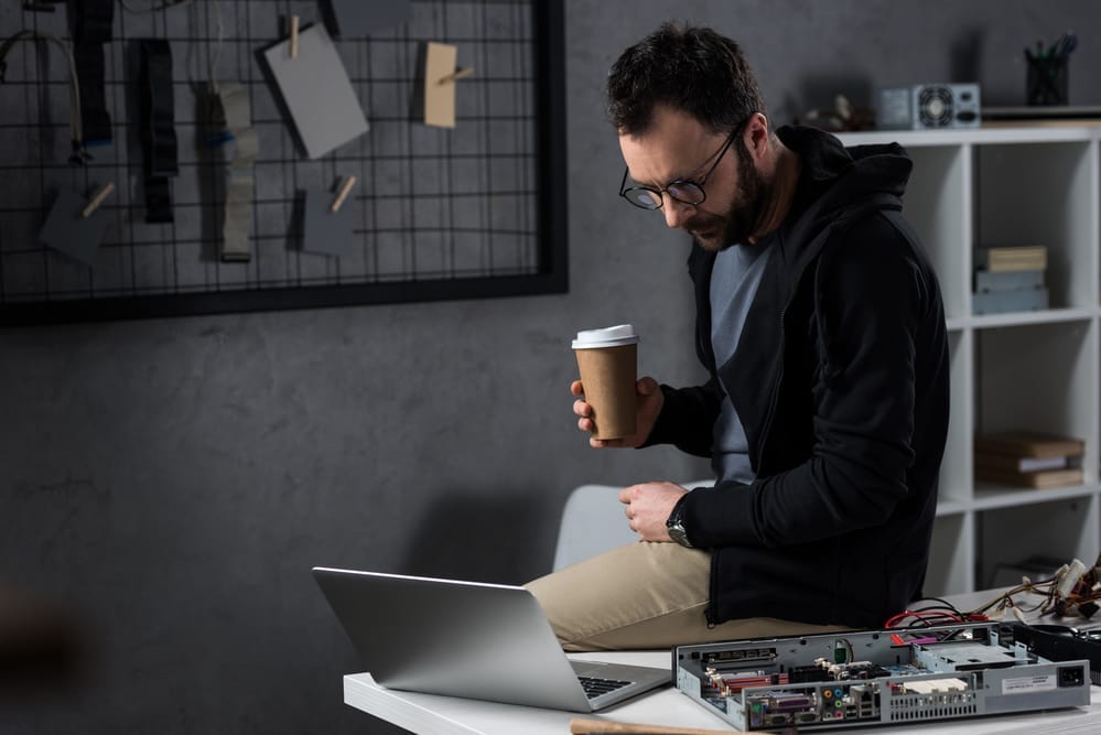 Man With Coffee In Hand Sitting On Table And Looking On Laptop